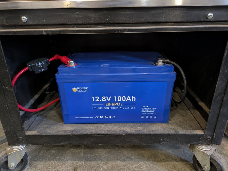 12.8V 100Ah LiFePO4 battery secured in bottom shelf of rolling cart with inline fuse on positive cable